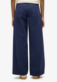 Vaqueros de pierna ancha azul marino con una cintura alta, dos bolsillos traseros y un acabado texturizado suave, combinados con zapatillas beige.