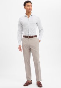 Camicia bianca con bottoni con motivo geometrico, pantaloni grigio chiaro e cintura e scarpe in pelle marrone, caratterizzati da un design aderente.