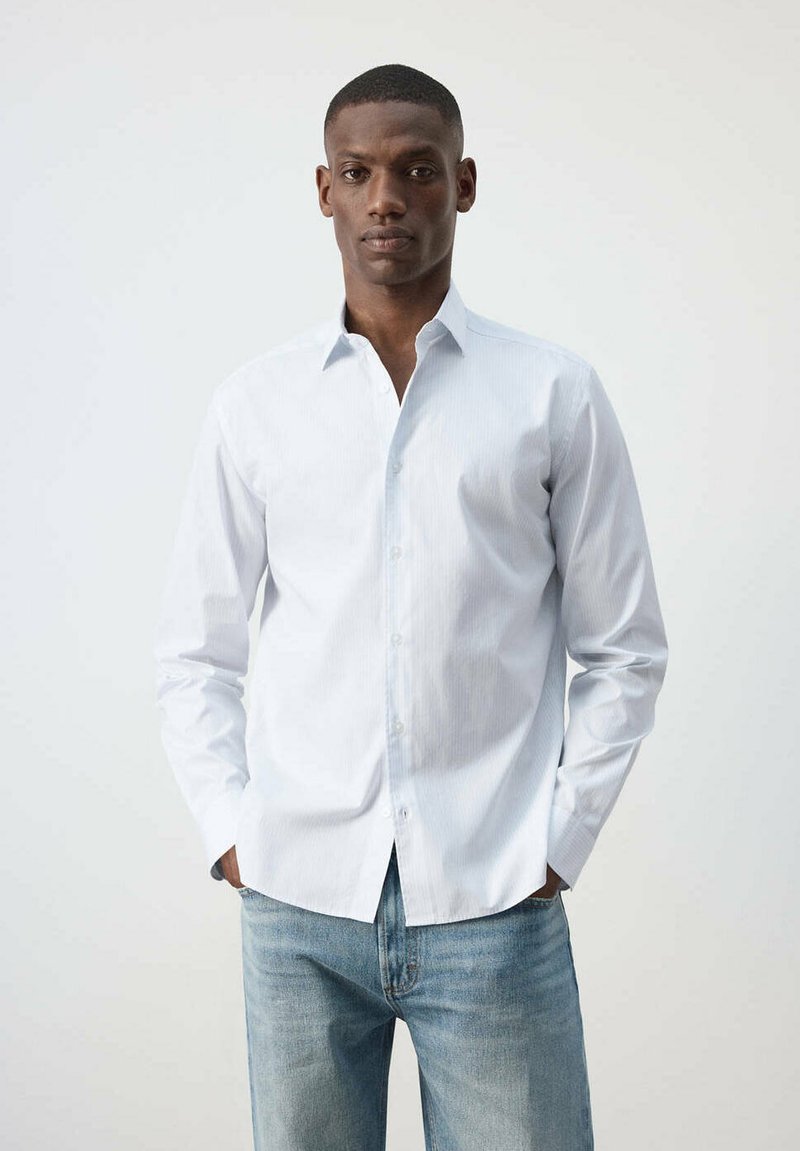 Homme debout, les mains dans les poches, portant une chemise blanche boutonnée et un jean bleu clair sur un fond blanc uni.