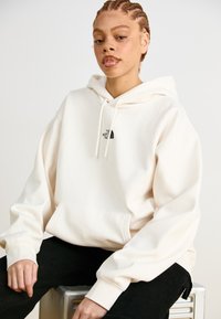 Felpa oversized color crema con tasca frontale e lacci bianchi, caratterizzata da un piccolo logo nero sul petto. Sotto si indossano pantaloni neri.