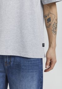 Gray cotton t-shirt with short sleeves, relaxed fit, and a black tag on the side. Worn with blue denim jeans, showing slight fading.