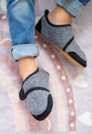 Grau gefütterte Hausschuhe mit schwarzen Akzenten, gesichert mit einem Riemen, haben eine abgerundete Zehe und eine sandfarbene Gummisohle. Getragen an mit Denim bekleideten Füßen.