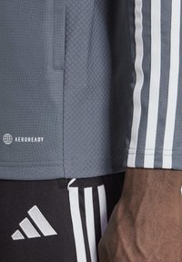 Gros plan sur un bras et une taille portant une veste Adidas grise et un pantalon Adidas noir, tous deux ornés d'un motif à trois bandes blanches et de logos.