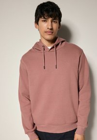 Sudadera de canalé rosa con cordón, cuenta con un bolsillo delantero y puños elásticos. Lleva puesta sobre una camisa blanca y se combina con pantalones oscuros.