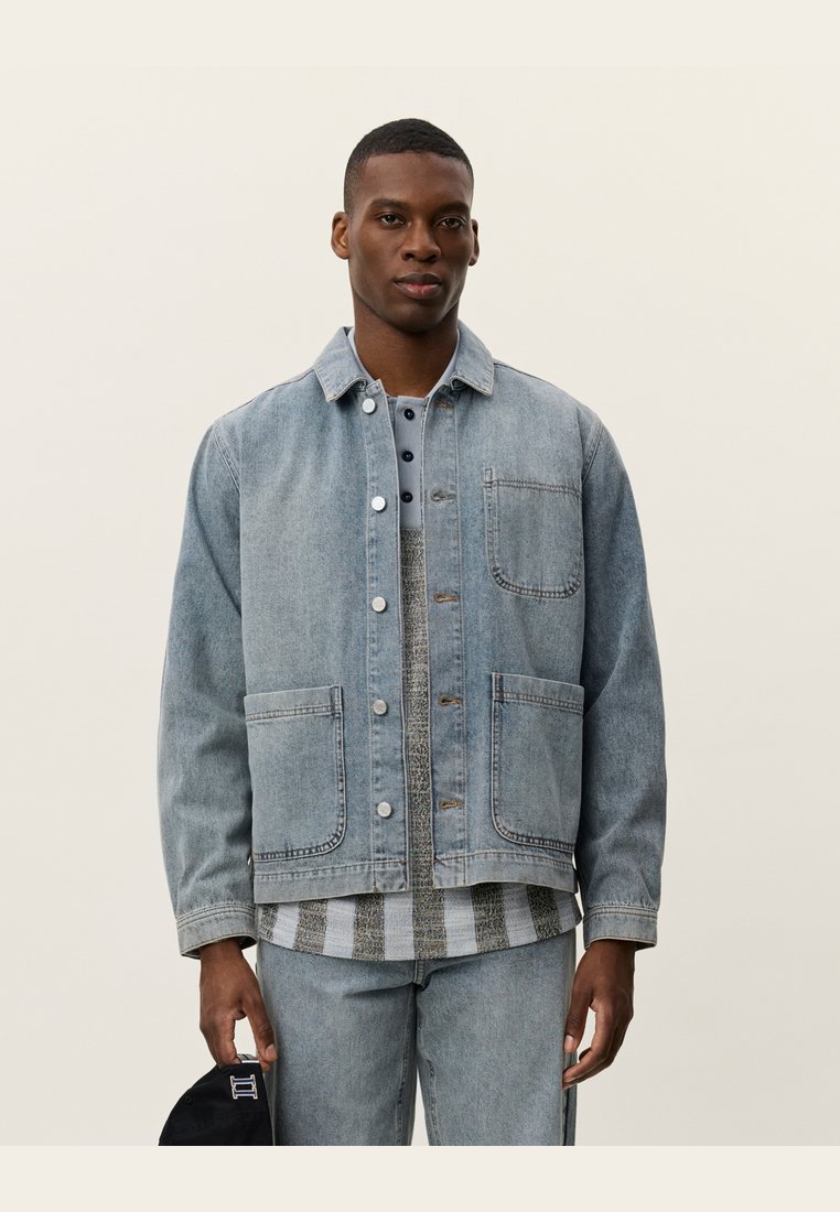 Denim jacket in light blue, featuring a button front, a collar, two front pockets, and a relaxed fit over a patterned shirt and jeans.