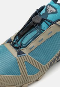Chaussure de sport turquoise et beige avec une tige en mesh texturé, des lacets élastiques et des accents en caoutchouc renforcé sur la semelle et les côtés.