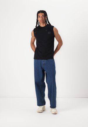 Svart ermeløs T-skjorte laget av bomull, kombinert med løstsittende blå denimjeans. Hvite teksturerte sneakers fullfører antrekket.