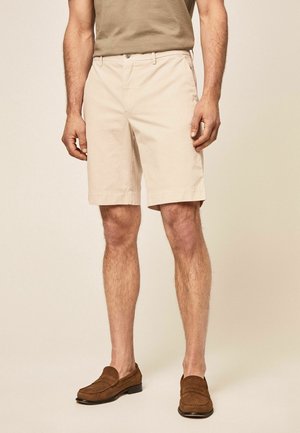 Homme portant un short beige aux genoux, des mocassins marron et une chemise marron à manches courtes, debout devant un fond beige.