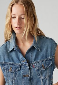 Gilet en denim bleu clair avec un col structuré, deux poches poitrine et des boutons métalliques marron. Tissu texturé avec des coutures visibles.