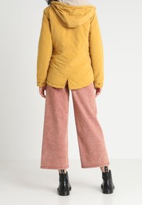 Żółta kurtka z kapturem o miękkim wnętrzu, w połączeniu z szerokimi różowymi spodniami z zastosowaniem korduroy i czarnymi, masywnymi butami, widoczna z tyłu.