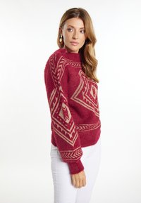 Pulover roșu, pufos, cu modele geometrice aurii, guler înalt și mansete ribbrate. Asortat cu pantaloni albi pentru contrast.