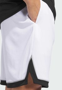 Weiße Sportshorts aus strukturiertem Mesh-Stoff, mit schwarzem Rand und seitlichen Schlitzen für bessere Bewegungsfreiheit. Sichtbares Taschendetail.