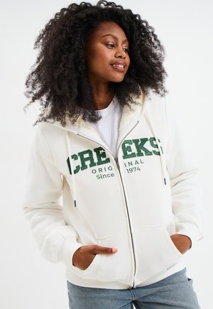 Veste à capuche zippée blanche avec le texte "CREEKS" en vert, des cordons de serrage et des poches avant. Matériau doux et capuche doublée d'une texture en polaire.