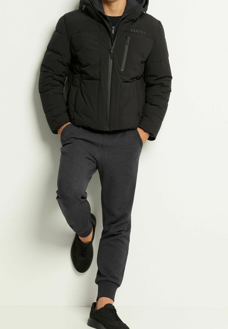 Homme portant une doudoune à capuche noire, un pantalon de jogging gris foncé et des baskets noires, debout contre un mur blanc les mains dans les poches.