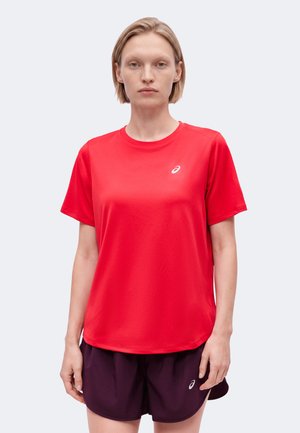 Jeune personne aux cheveux courts et blonds, portant un t-shirt de sport rouge à manches courtes et un short violet foncé, debout devant un fond uni.