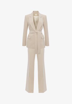 Beżowa marynarka dopasowana z paskiem w talii i dwoma przednimi kieszeniami, zestawiona z szerokimi spodniami w pasującym kolorze i tkaninie. Gładka tekstura.