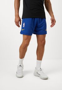 Kék hálós kosárlabadanadrág fehér NBA logóval, sárga Nike pipa és „Warriors” felirattal. Fehér sportcipővel és zoknikkal párosítva.