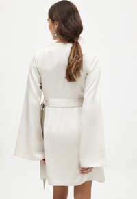 Robe crème, arrivant au genou, en satin, avec des manches évasées et un détail de taille noué. Texture lisse avec un design minimaliste.