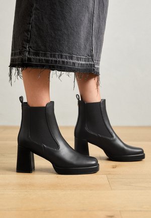 Bottes chevilles en cuir noir avec panneaux latéraux élastiques, bout carré et talons blocs épais. Portées avec une jupe en denim gris à ourlet effiloché.