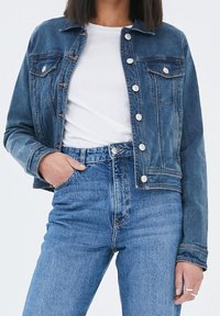 Veste en denim bleu foncé avec un devant à boutons, deux poches poitrine et des accents en métal argenté. Portée par-dessus un t-shirt blanc et associée à un jean bleu.