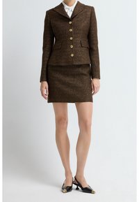 Femme portant un blazer marron texturé boutonné avec une jupe courte assortie et des chaussures noires à talons bas avec des détails dorés.