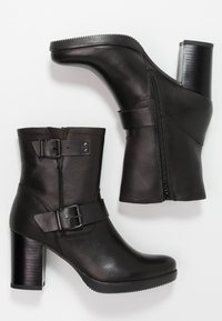 Botas de cuero negras hasta el tobillo con un tacón grueso. Las características incluyen una cremallera lateral, correas ajustables con hebilla y una textura suave y brillante.