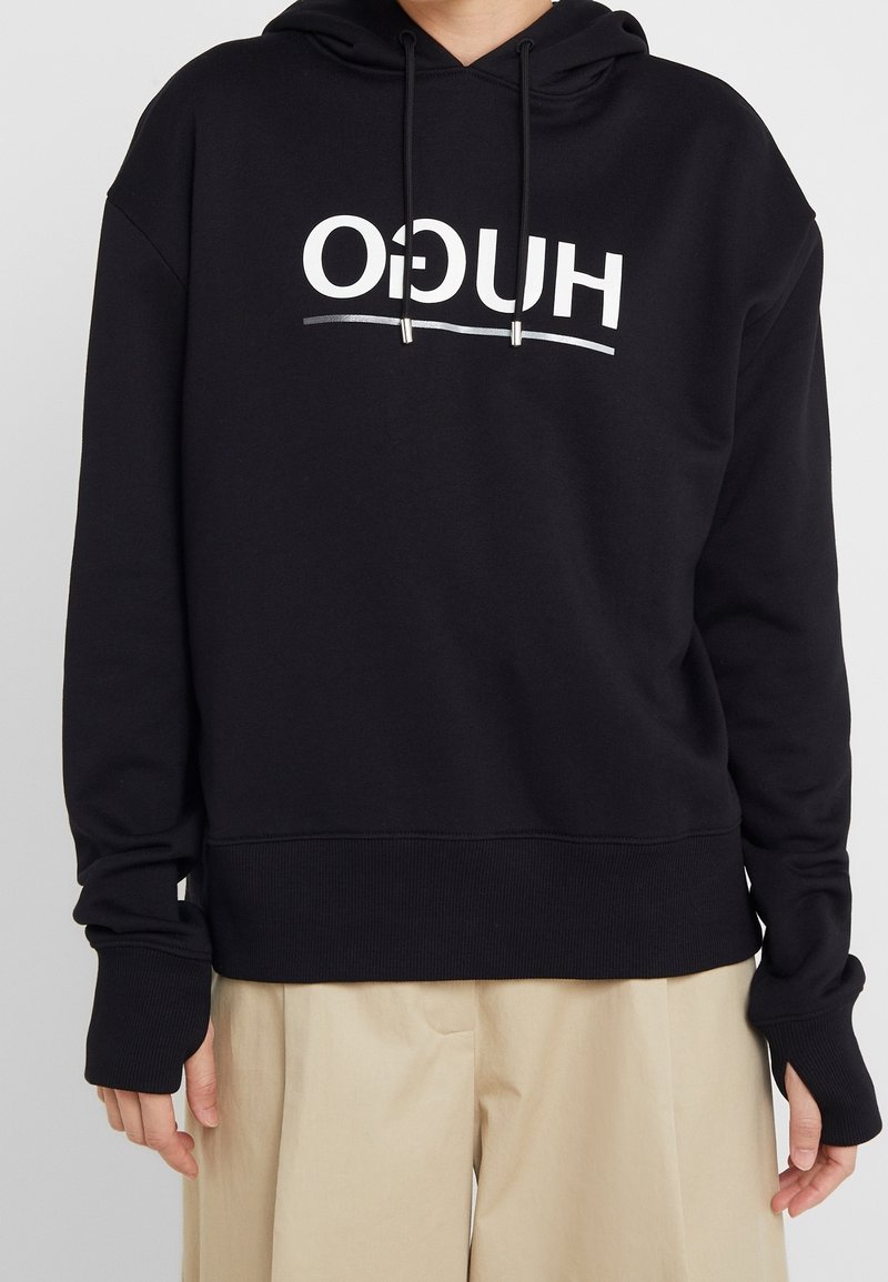 Person som bär en svart hoodie med vit text "HUGO" upp och ner och beige byxor, stående mot en enkel ljus bakgrund.