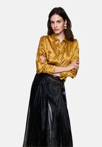 Golden satin blouse with a textured pattern; long black pleated skirt featuring a subtle sheen and a chain detail at the waist.