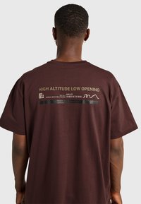Man med kort hår och guldfärgade ringörhängen som bär en brun T-shirt med texten "HIGH ALTITUDE LOW OPENING" och grafik på baksidan.