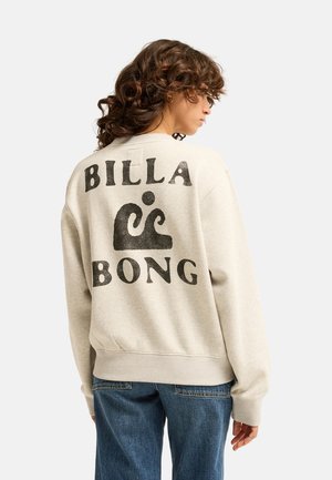 Kobieta z kręconymi włosami, ubrana w jasnobeżową bluzę z czarnym logo "Billabong" na plecach oraz niebieskie dżinsy, zwrócona bokiem.