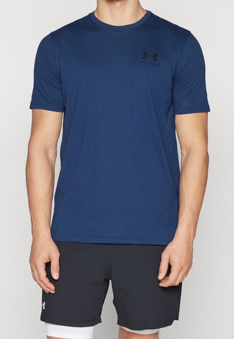 Muž má na sobě tmavě modré tričko s krátkým rukávem Under Armour a černé sportovní šortky s bílými kompresními legínami pod nimi.