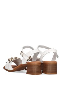Sandalias de cuero blanco con un pequeño tacón cuadrado marrón, que presentan un acento decorativo de flor y correas ajustables en el tobillo. Suela texturizada.