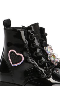 Botas de tobillo de charol negro con un parche en forma de corazón, acentos de piedras preciosas y cordones negros. Presentan un acabado brillante destacado y una pestaña para tirar.
