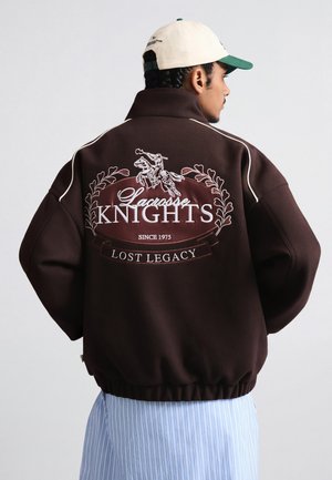 Person trägt eine dunkelbraune Jacke mit der Stickerei "Lacrosse Knights Lost Legacy Seit 1975" und eine beige Baseballkappe, seitlich vom Betrachter gewandt.