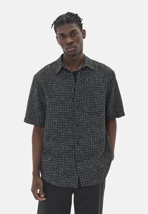 Chemise à manches courtes noir et blanc avec un motif à carreaux, présentant un design à boutons et une coupe décontractée. Portée par-dessus une chemise noire.