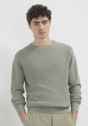 Hellgrüner gerippter Strickpullover mit Rundhalsausschnitt und langen Ärmeln, der über einen strukturierten Stoff und schmale Bündchen verfügt. Kombiniert mit beigen Hosen.
