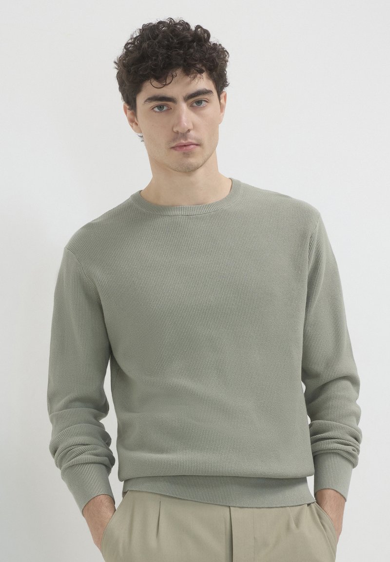 Pull en maille côtelée vert clair avec un col rond et des manches longues, présentant un tissu texturé et des poignets ajustés. Associé à un pantalon beige.