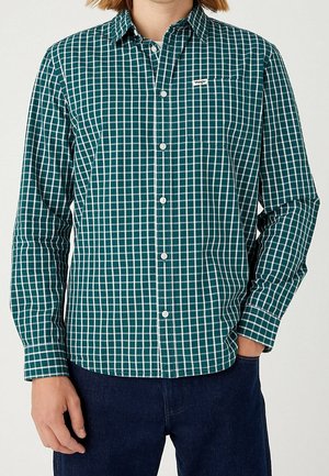 Homme portant une chemise à carreaux verts et blancs à manches longues et un pantalon bleu foncé, debout avec les mains baissées.