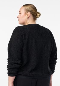 Sieviete ar blondiem matiem, kas savīti bunā, valkā sudraba riņķa auskarus un melnu teksturētu garām piedurknēm flīsa jaku, stāv profila pozā pret baltu fona.