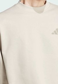Světle béžová mikina s kulatým výstřihem, měkkou texturou, se slabým šedým logem Adidas na levé straně hrudi.