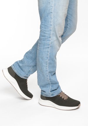 Person trägt hellblaue Jeans und schwarze Sportschuhe mit weißen Sohlen, ein Fuß leicht hinter dem anderen angehoben.