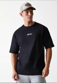 T-shirt preta de algodão com gola redonda e mangas curtas, com o logótipo "New Era" branco no peito. O modelo também usa um boné de basebol cinzento.