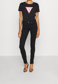 Kvinde iført sort Guess t-shirt, sorte tætsiddende jeans og sorte højhælede sandaler står foran en ensfarvet lys baggrund.