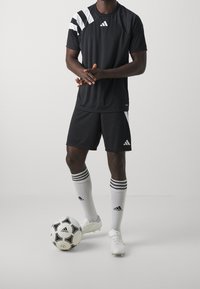 Černé atletické šortky a tričko s bílými pruhy a logem, doplněné bílými kopačkami a vysokými ponožkami s třemi pruhy. Černobílý fotbalový míč.