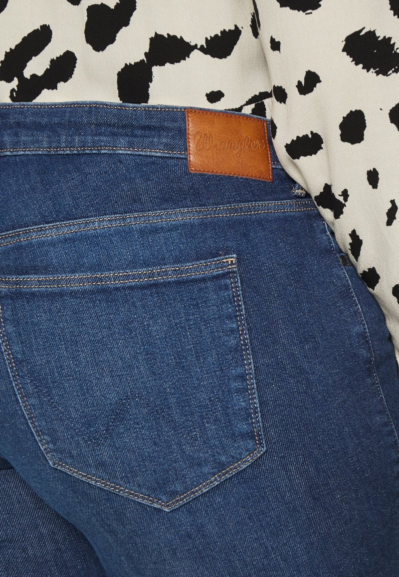 Gros plan sur la poche d’un jean en denim bleu avec une étiquette en cuir de la marque Wrangler, portée avec un haut en tissu à pois noirs et blancs.