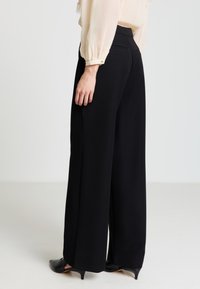 Pantaloni neri a gamba larga con una texture liscia, vita strutturata e due tasche posteriori, abbinati a décolleté nere a punta.