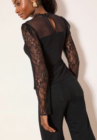 Fekete csipketop hosszú, áttetsző csipke ujjakkal, testhez simuló felsőrész és kulcslyuk alakú hátbeli részlettel, fekete nadrággal párosítva. Texturált csipkeminta.