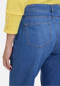Blauwe denim jeans met een aansluitende taille, voorzien van een achterzak en een gladde textuur. Een gele, gestructureerde trui met knoopaccenten.