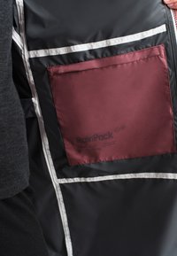 Osoba u crnoj jakni s crnim unutarnjim podstavom i bijelim šavovima, te velikim tamnocrvenim džepom označenim "RainPack."