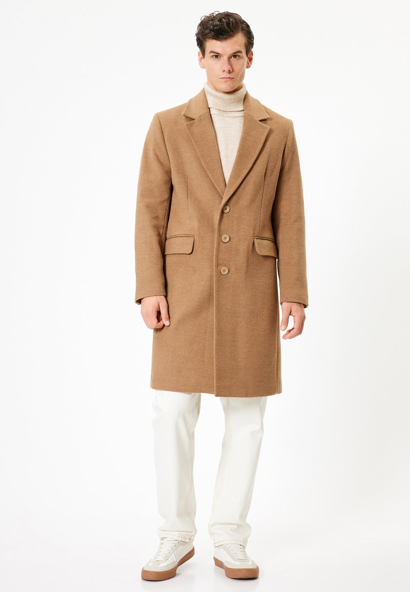 Cappotto in misto lana color tan con revers, due tasche frontali e tre bottoni. Indossato sopra un dolcevita beige chiaro e abbinato a jeans bianchi.